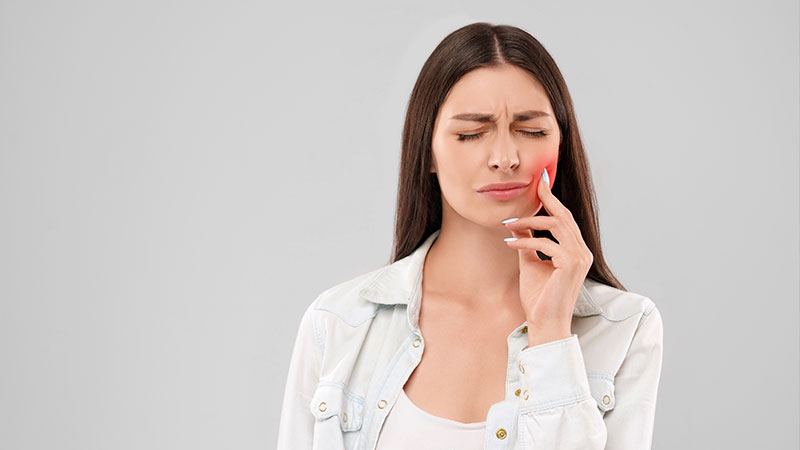 uma mulher branca, de cabelos castanhos longos e lisos, está com um semblante de dor e desconforto. Com a mão esquerda, ela pressiona a bochecha do mesmo lado, que está vermelha. A mulher usa uma jaqueta jeans e uma camiseta branca. O fundo da imagem é cinza.