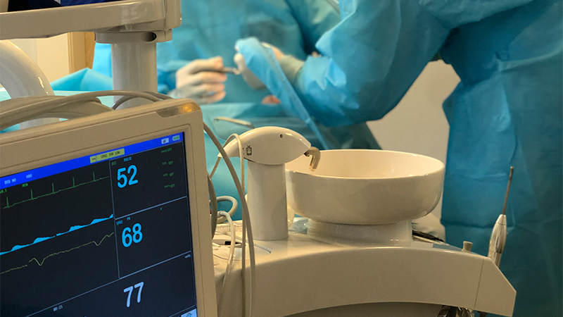 No canto esquerdo da imagem há um medidor automático de pressão arterial e batimentos e ao fundo dois dentistas com roupas azuis realizando uma operação.