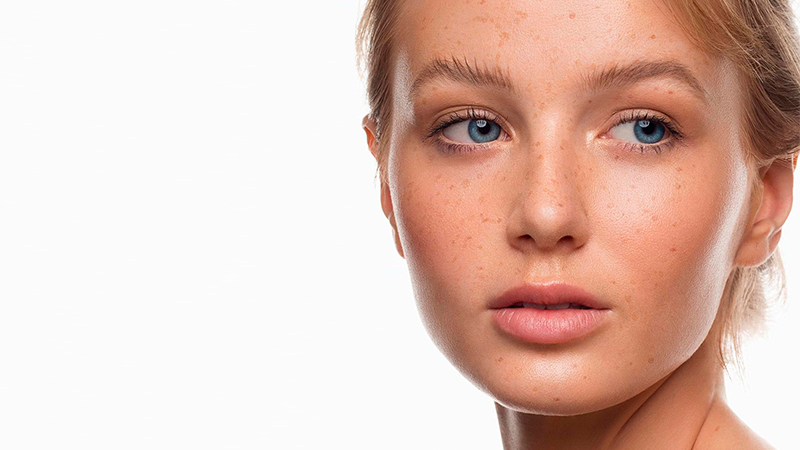 Mulher branca, de cabelo loiro, no centro da imagem, com fundo rosa-claro. A imagem exibe o terço superior do corpo da mulher, dos ombros até a cabeça. Ela está com o cabelo preso, posicionada de lado, virada para a direita, olhando para baixo, com a mão esquerda tocando levemente o seu rosto.