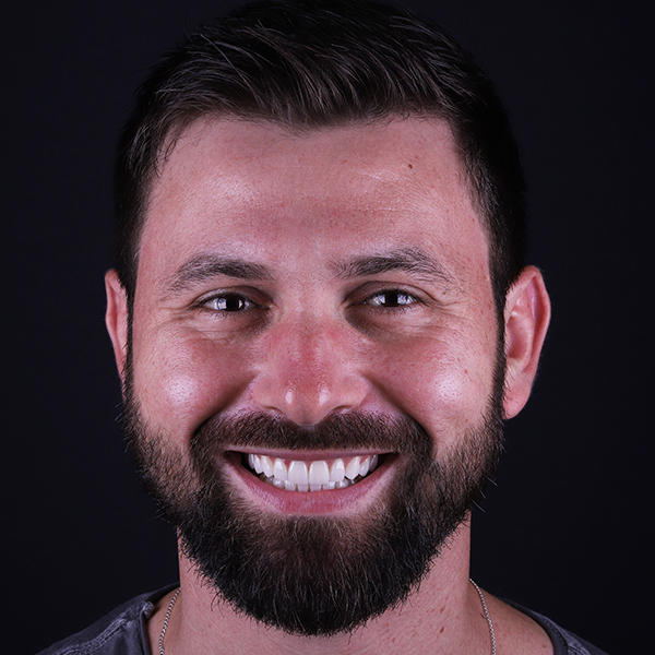 Um homem branco, de cabelos e barba grossos e escuros, sorri para a câmera mostrando os dentes. Ele está em frente a um fundo preto e só é possível ver sua cabeça e pescoço.