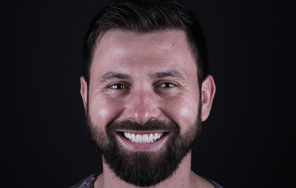 um homem branco, de olhos castanhos e cabelos escuros, sorri. O fundo da imagem é preto.