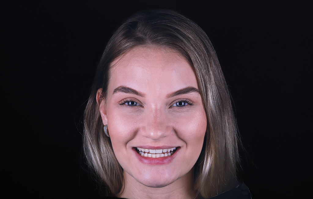 uma mulher branca, loira e de olhos claros sorri. Ela está com os cabelos lisos soltos. O fundo da imagem é preto.