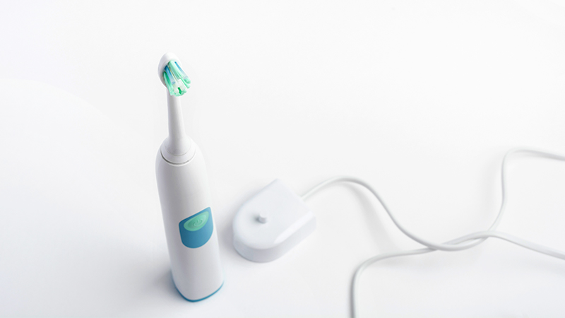 No centro, mais embaixo da imagem, está uma escova de dente elétrica branca, com detalhes azuis e verdes nas cerdas. Ao seu lado está um carregador quadrado e branco com um fio. O fundo da imagem é branco.