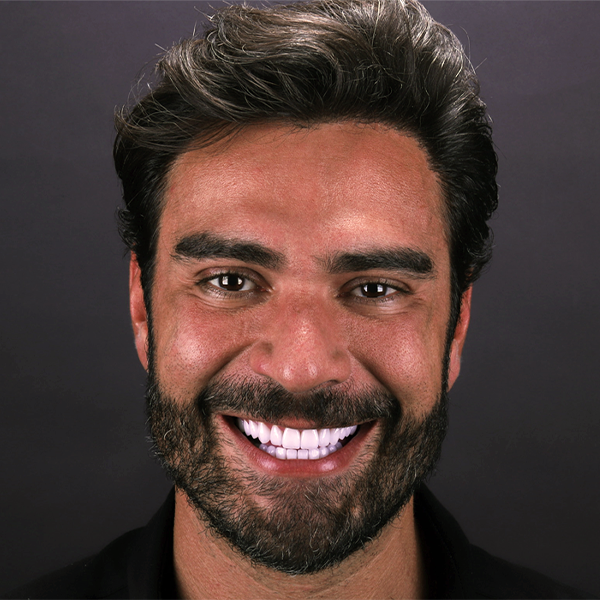 Imagem em close do rosto de um homem branco, com cabelos, olhos e barba escuros. Ele sorri olhando para frente. O fundo da imagem é preto.