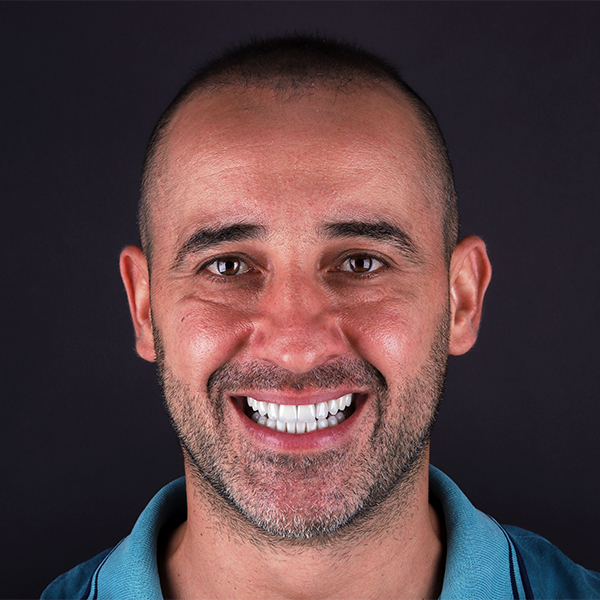 Imagem em close do rosto de um homem branco, com cabelo raspado. Ele usa uma camisa polo azul e sorri olhando para frente. O fundo da imagem é preto.