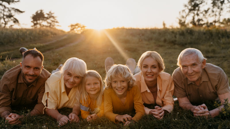 Imagem de uma família deitada de bruços sobre um campo durante o pôr do sol. Da esquerda para a direita estão: o pai, com cabelos e barba castanhos e uma camisa marrom; a avó, com cabelos brancos e uma camisa amarela; a filha criança, com cabelos loiros compridos e uma camiseta laranja; o filho pré-adolescente, com camiseta laranja e cabelos loiros; a mãe, com uma camisa salmão e cabelos loiros presos; e o avô, com cabelos brancos e polo marrom. Todos sorriem olhando para frente.